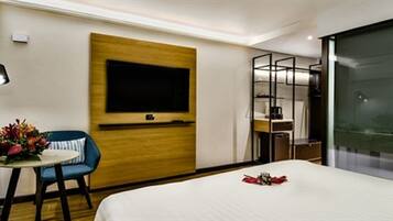 A hotel room with a bed, a desk with a chair, a TV mounted on the wall, and a glass partition.