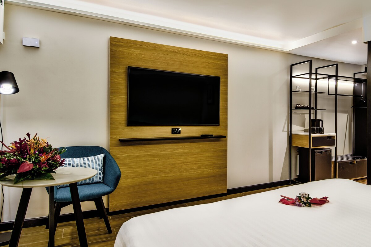 Habitación de hotel con cama, escritorio con silla, televisor montado en la pared, una pequeña mesa con arreglo floral y un panel de vidrio.