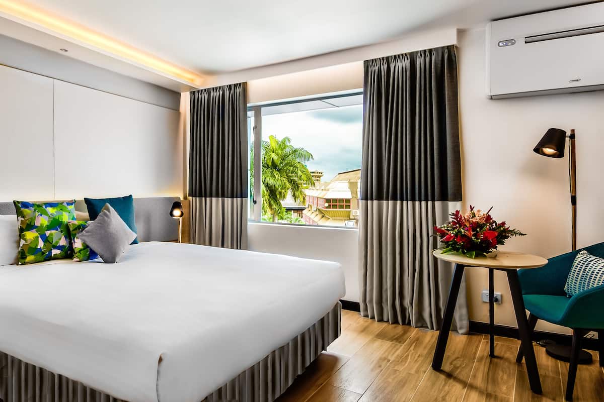 A hotel room with a large bed, a desk with a lamp, a small table with flowers, and a window with curtains.