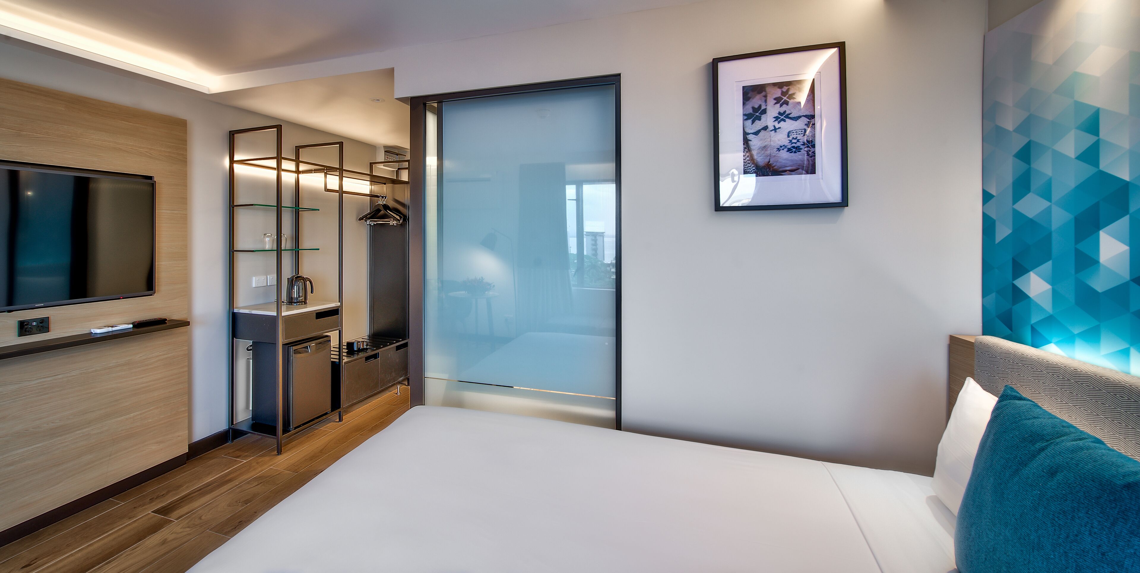 Een moderne hotelkamer met een groot flatscreen televisietoestel, een inloopkast, een glazen schuifdeur naar een balkon en een lijst met abstracte kunst.