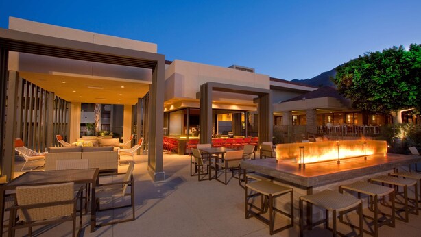 An outdoor dining area with tables and chairs, a fire feature, and a bar area with red chairs. - Hilton Palm Springs Resort (Palm Springs)