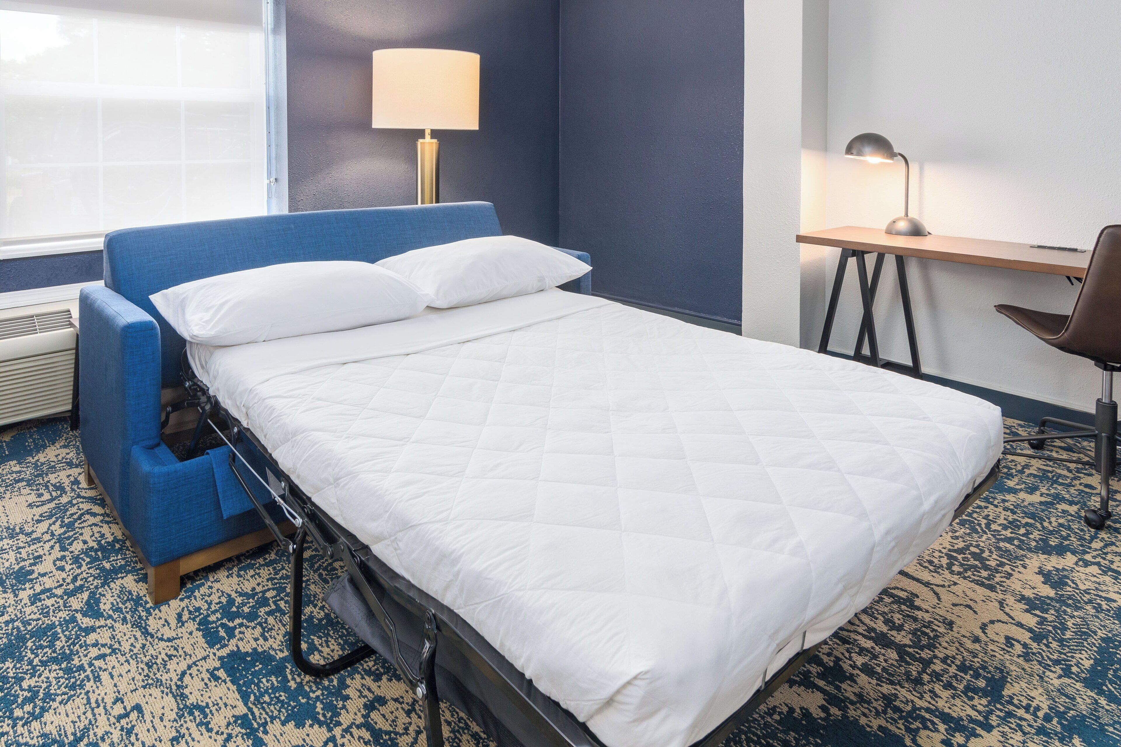 A hotel room with a bed, a desk, a chair, a lamp, and a window.