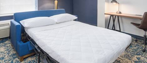 A hotel room with a bed, a desk, a chair, a lamp, and a window.