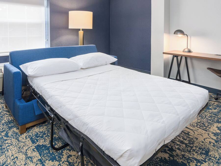 A hotel room with a bed, a desk, a chair, a lamp, and a window.