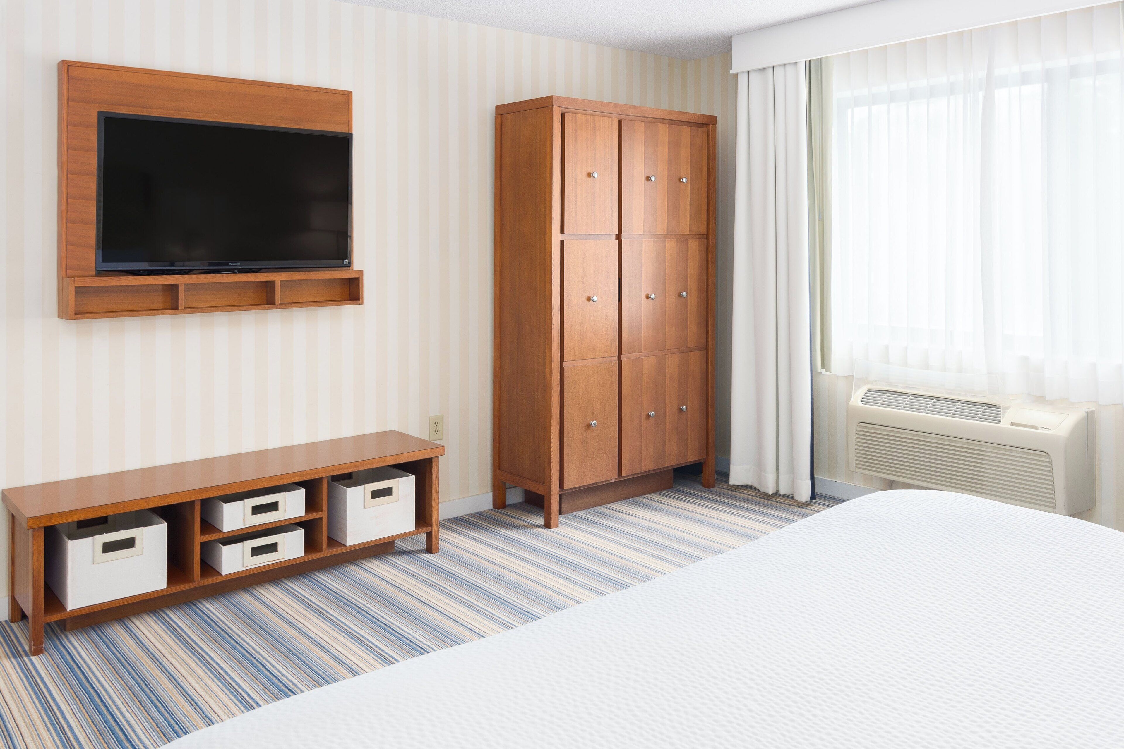 Habitación de hotel con una cama grande, un televisor montado en la pared, un armario de madera y una pequeña mesita con cajones.