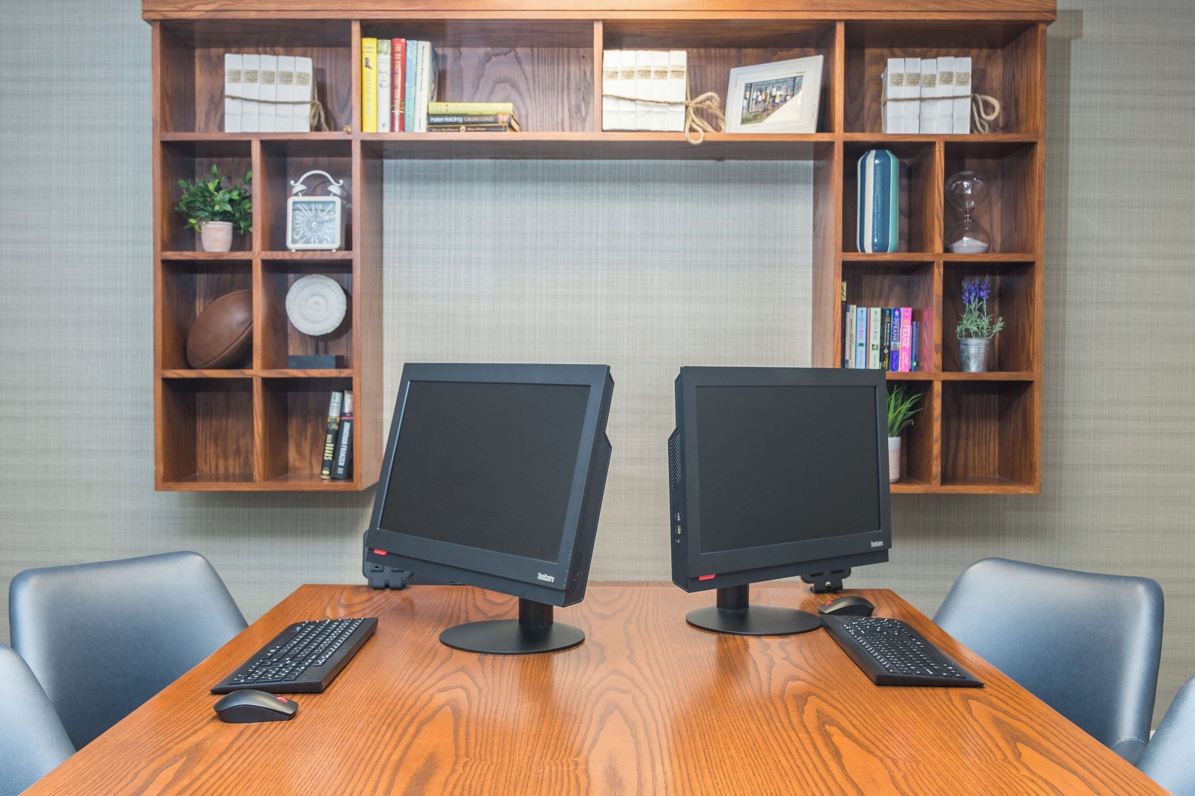 A workspace with two monitors, a wooden desk, and a bookshelf filled with books and decorative items.