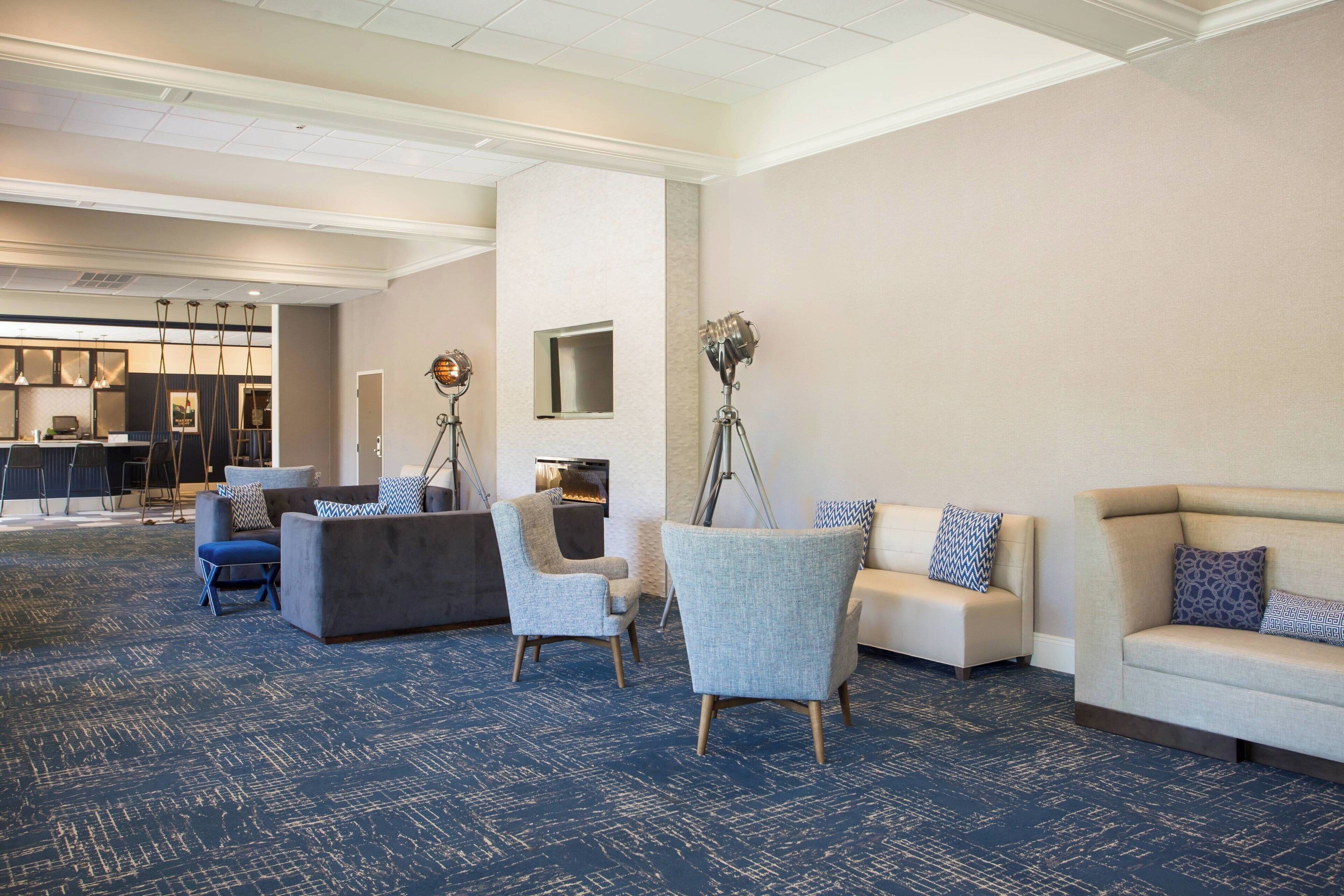 A spacious lobby with a blue carpet, multiple seating areas including sofas and armchairs, a fireplace, and a kitchen area in the background.