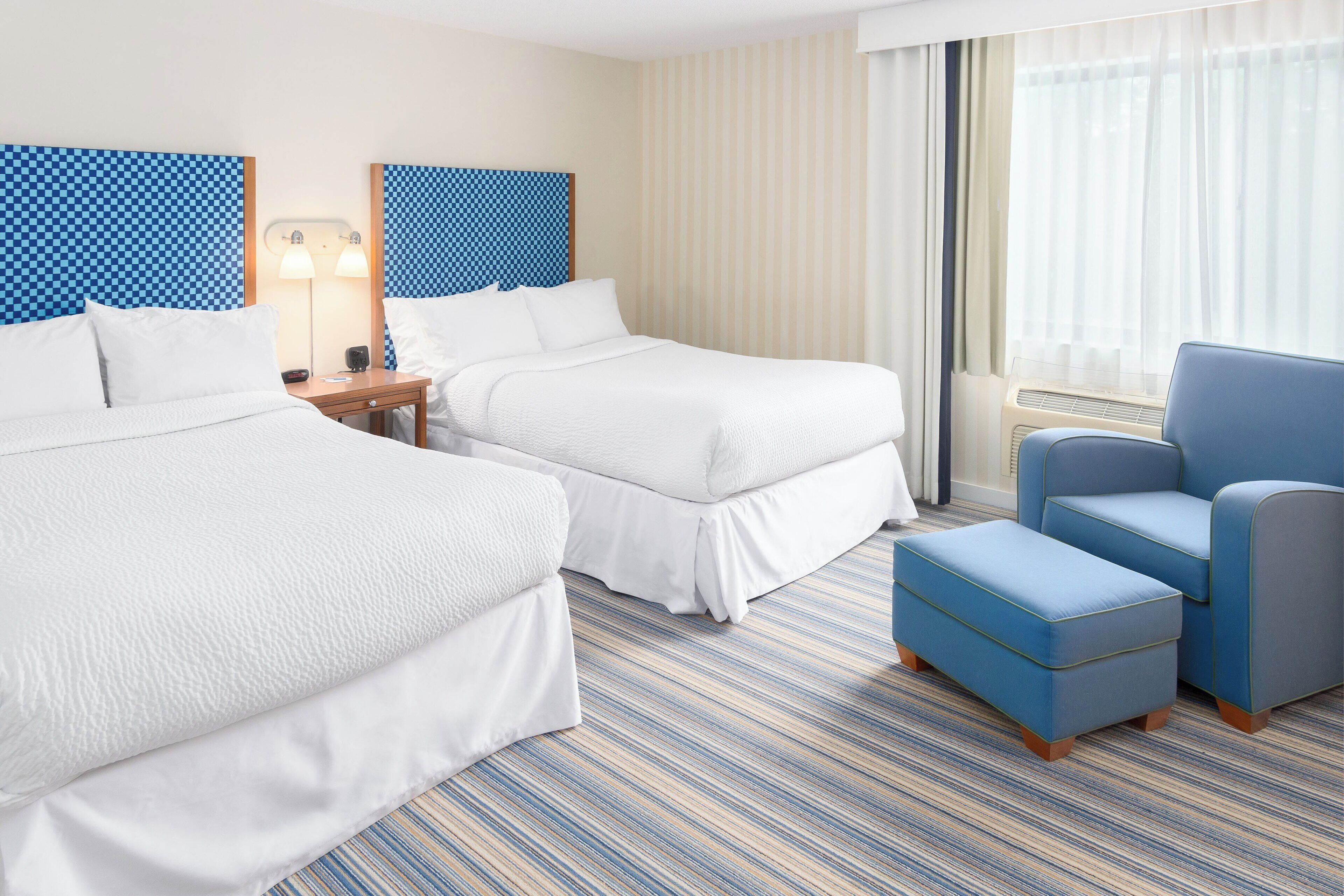 Habitación de hotel con dos camas, un escritorio con lámpara, un sillón azul y un amplio ventanal con cortinas.