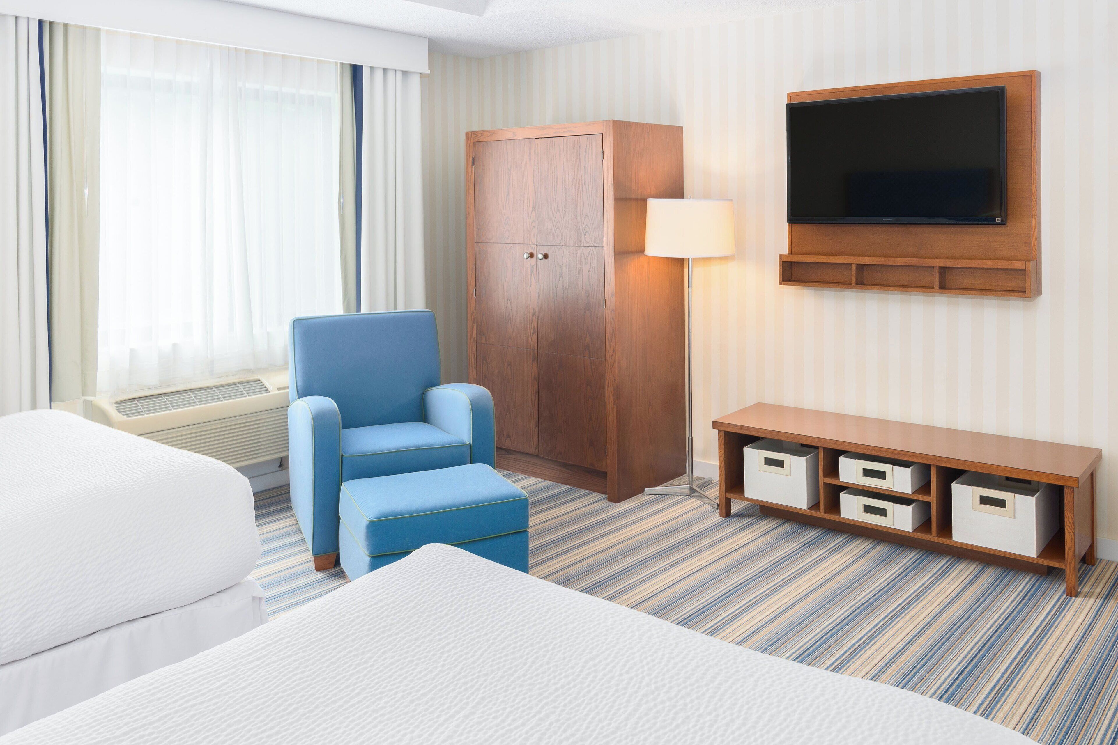 A hotel room with a bed, a blue armchair, a wooden wardrobe, a TV mounted on the wall, and a bedside table with drawers.