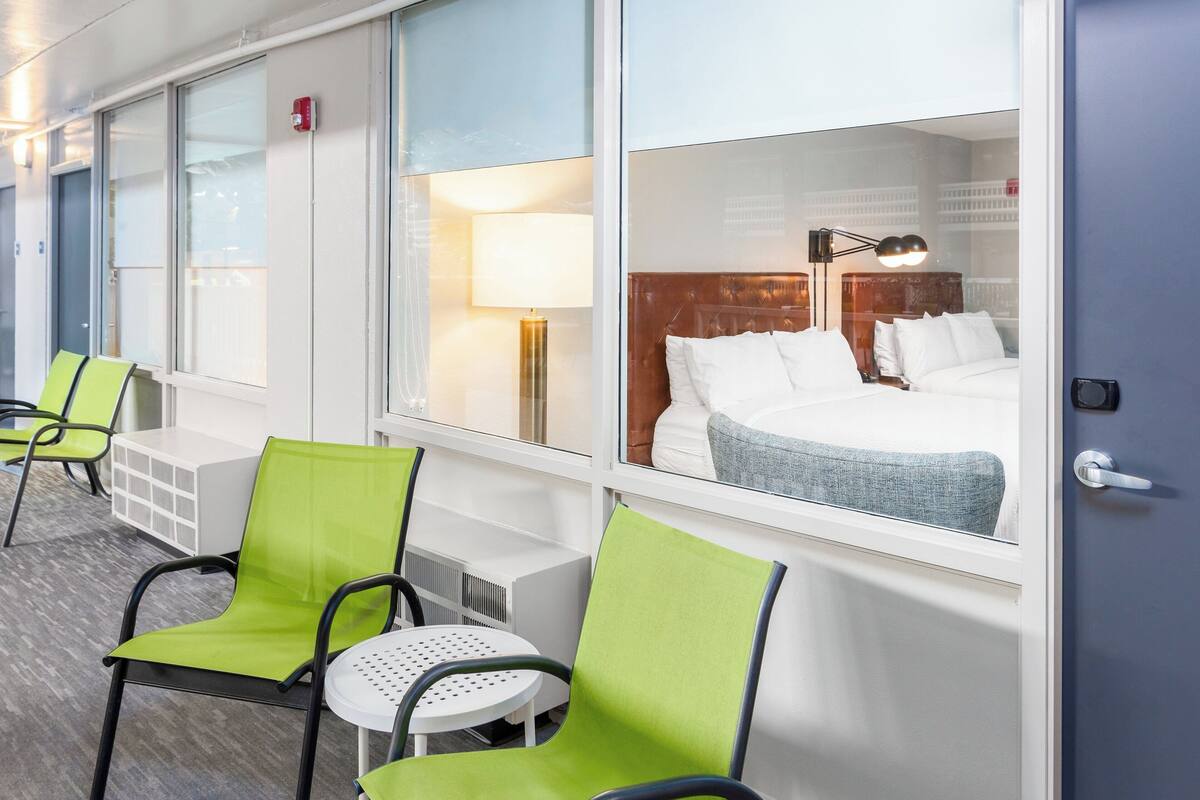 Habitación de hotel con cama, mesita de noche, lámpara y ventana con vista a la ciudad.
