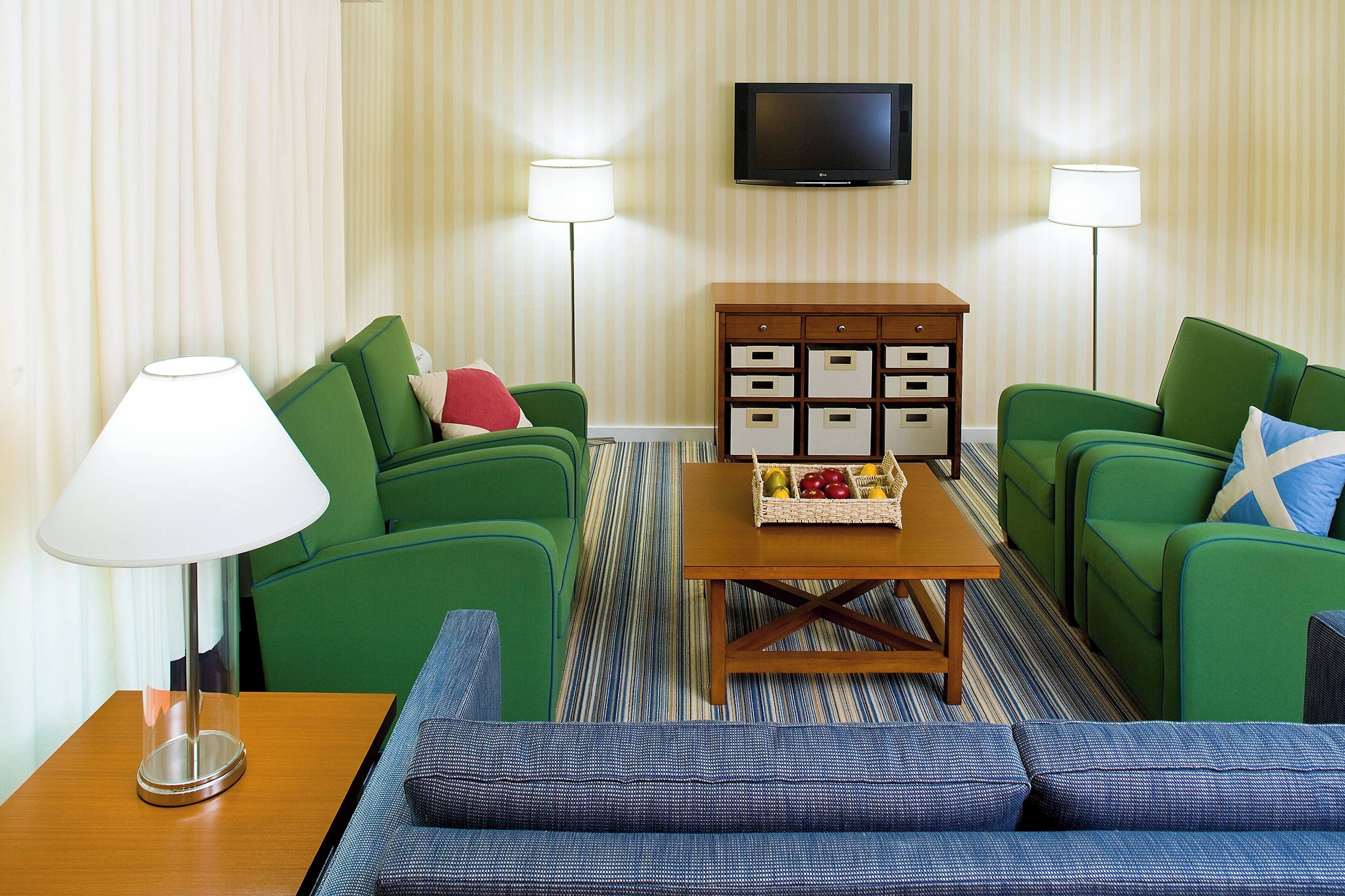 Een hotelkamer met twee groene banken, een houten bijzettafeltje met fruit, een televisie en twee vloerlampen.