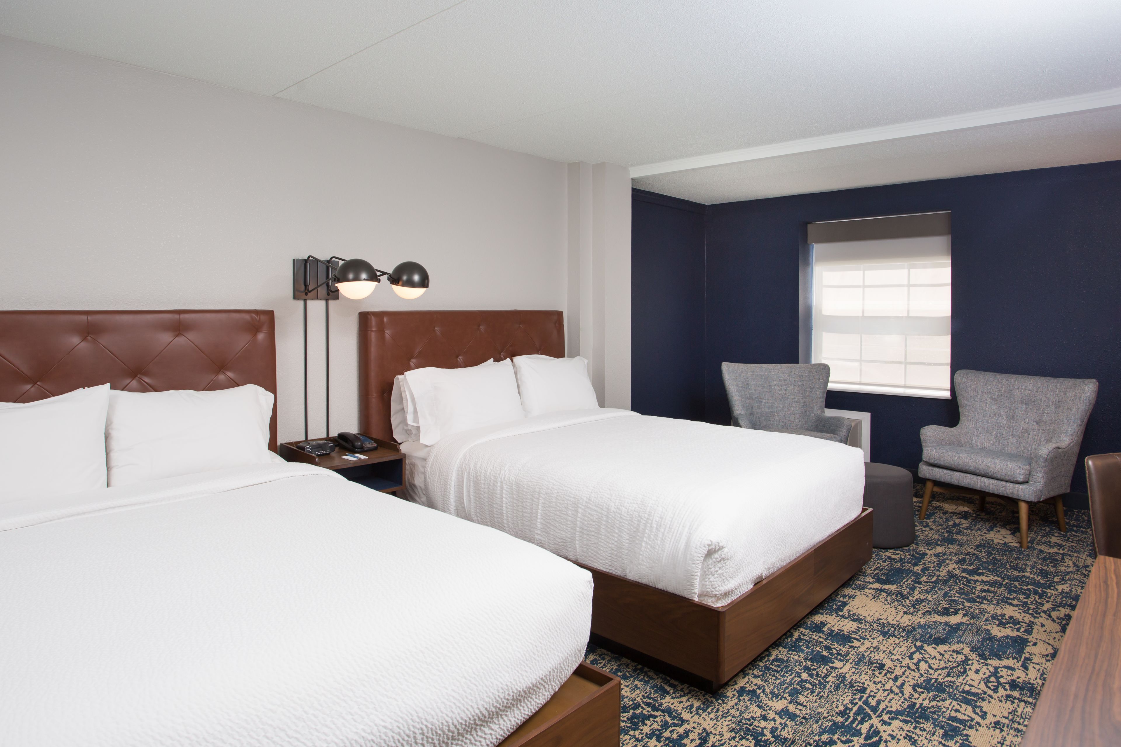 A hotel room with two beds, a nightstand, a lamp, a chair, and a window.