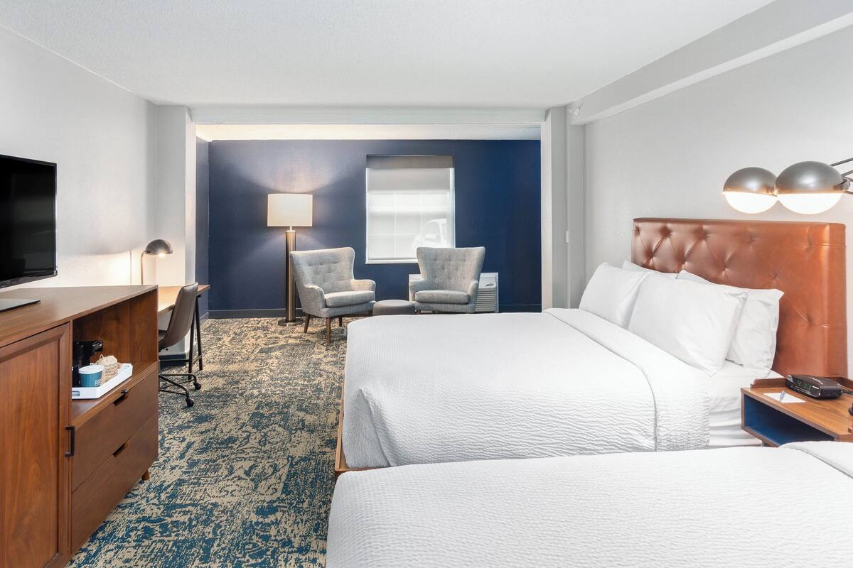 Habitación de hotel con una cama grande, un escritorio con lámpara, dos sillones, un televisor y una ventana con persianas.
