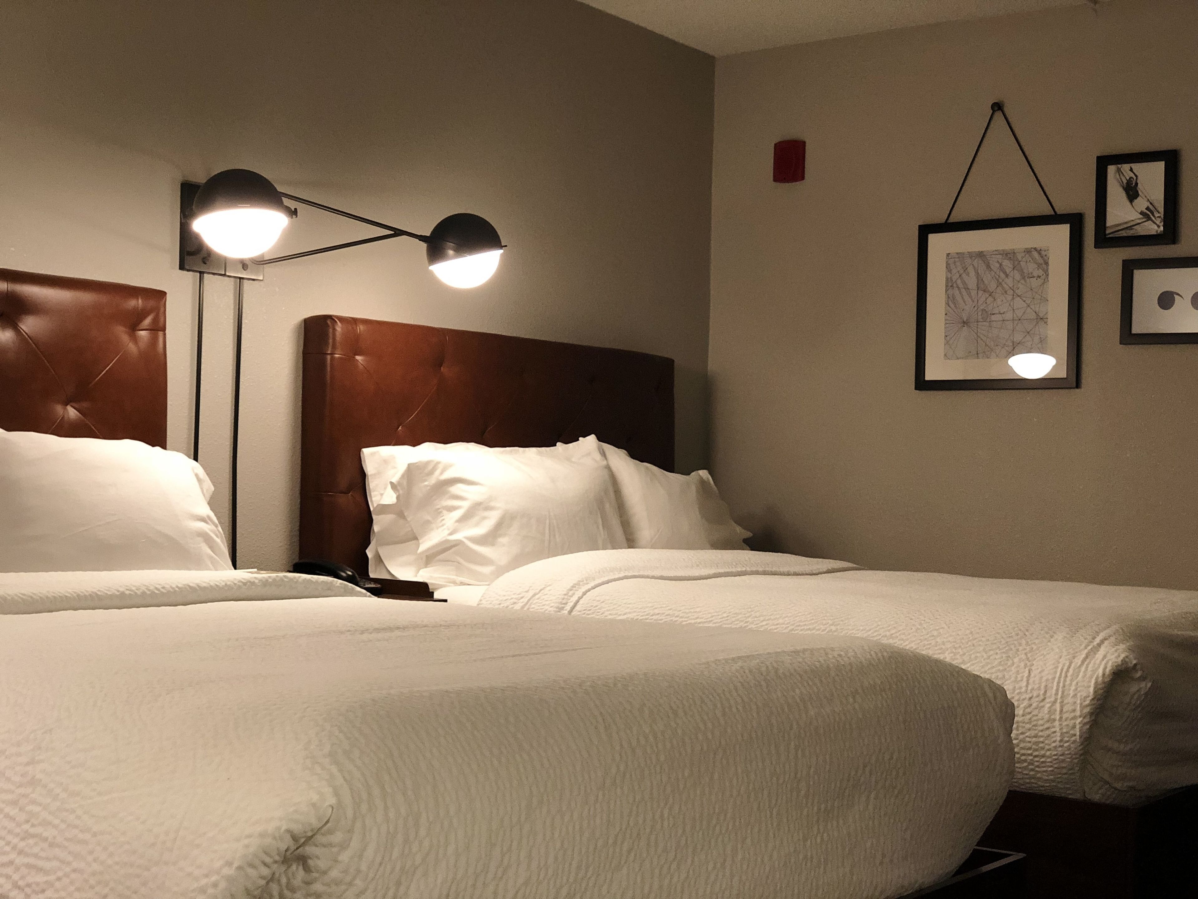 A hotel room with two beds, a brown headboard, white bedding, a floor lamp, and framed artwork on the wall.