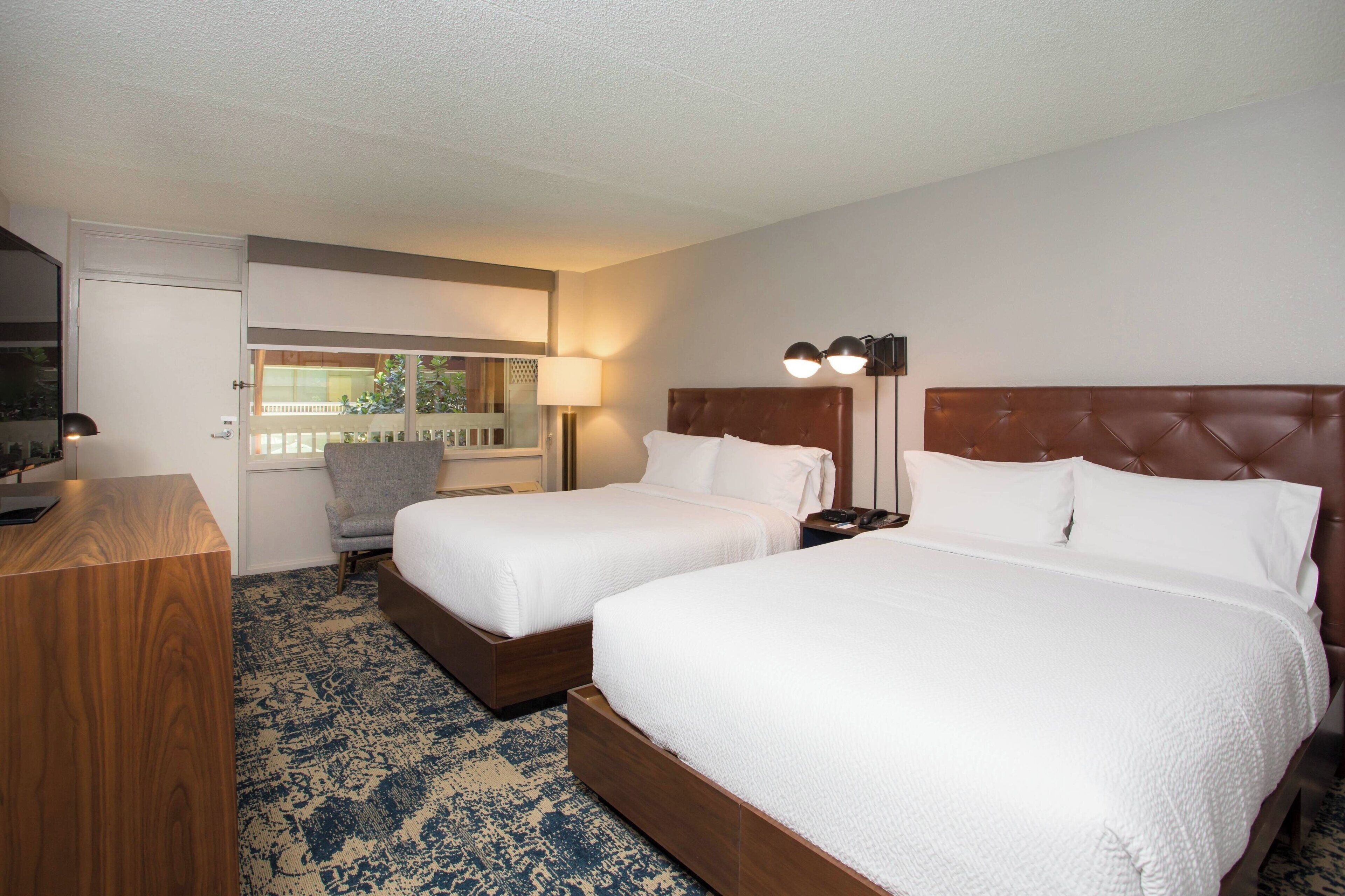 A hotel room with two beds, a desk, a chair, a TV, and a window with a view of greenery.