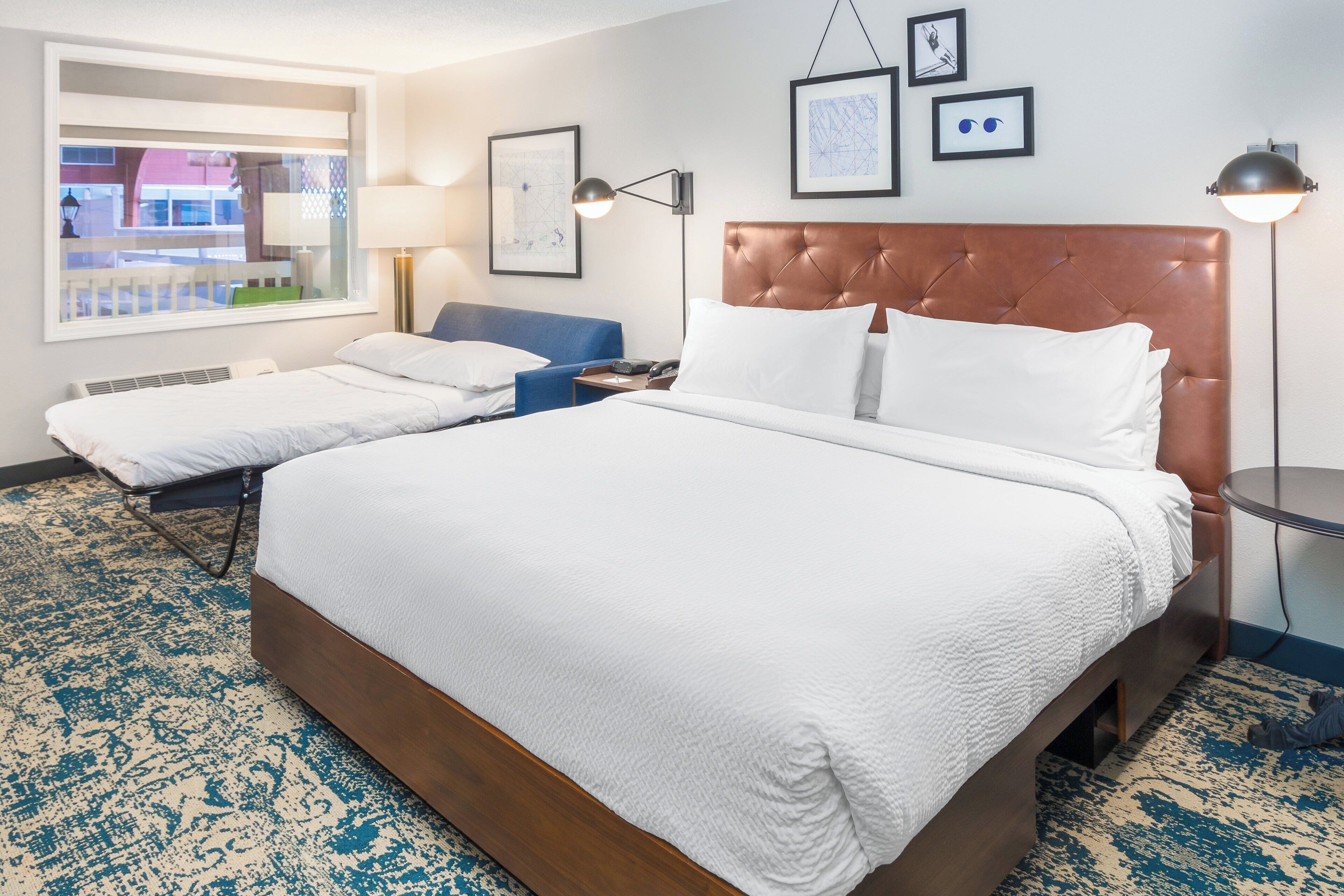 Ein Hotelzimmer mit einem großen Bett, einem kleineren Bett, einem Schreibtisch und einem Fenster mit Blick auf ein Gebäude.