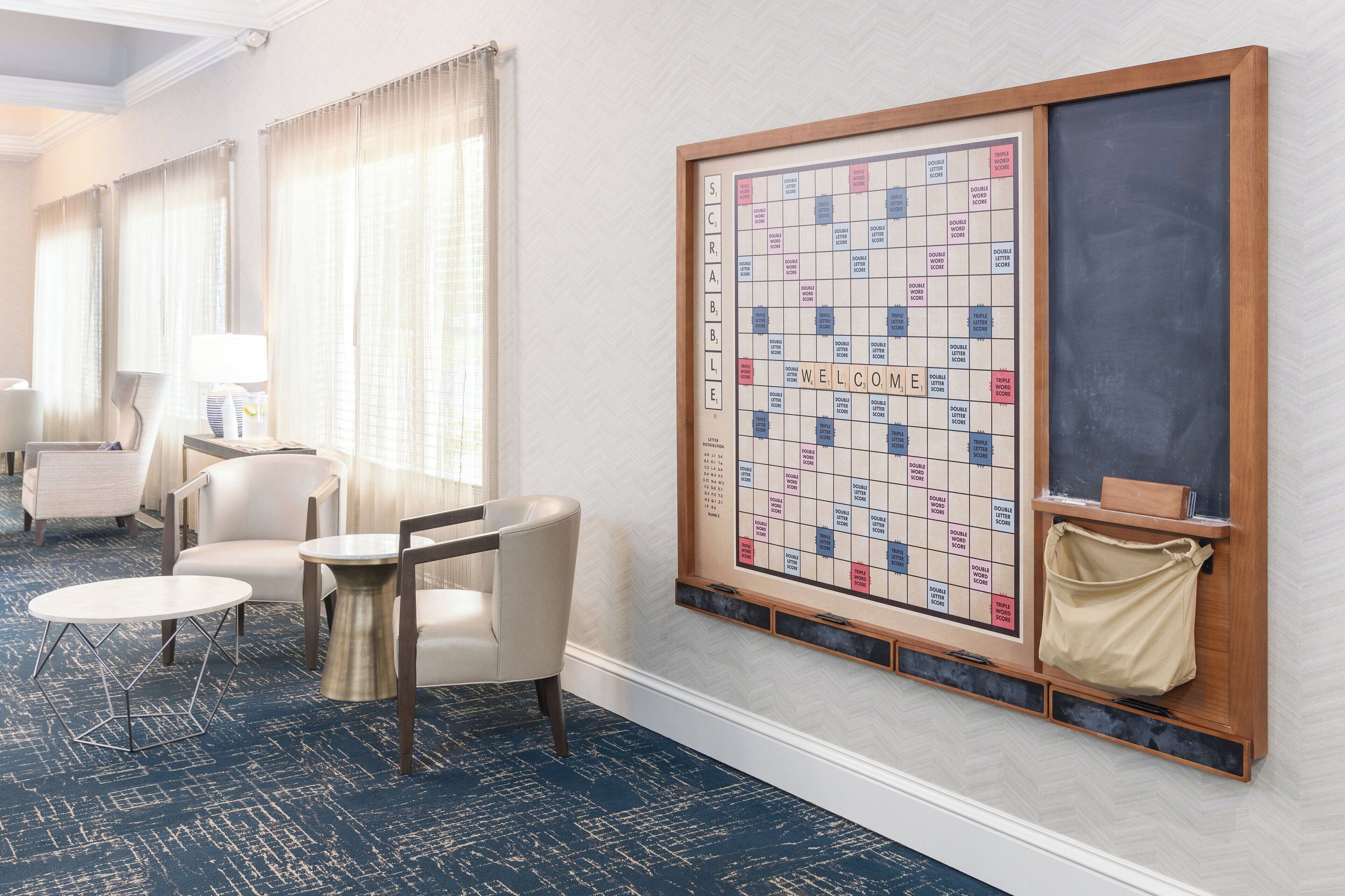 A hotel room with a large Scrabble board on the wall, a round table, two chairs, and a lamp.