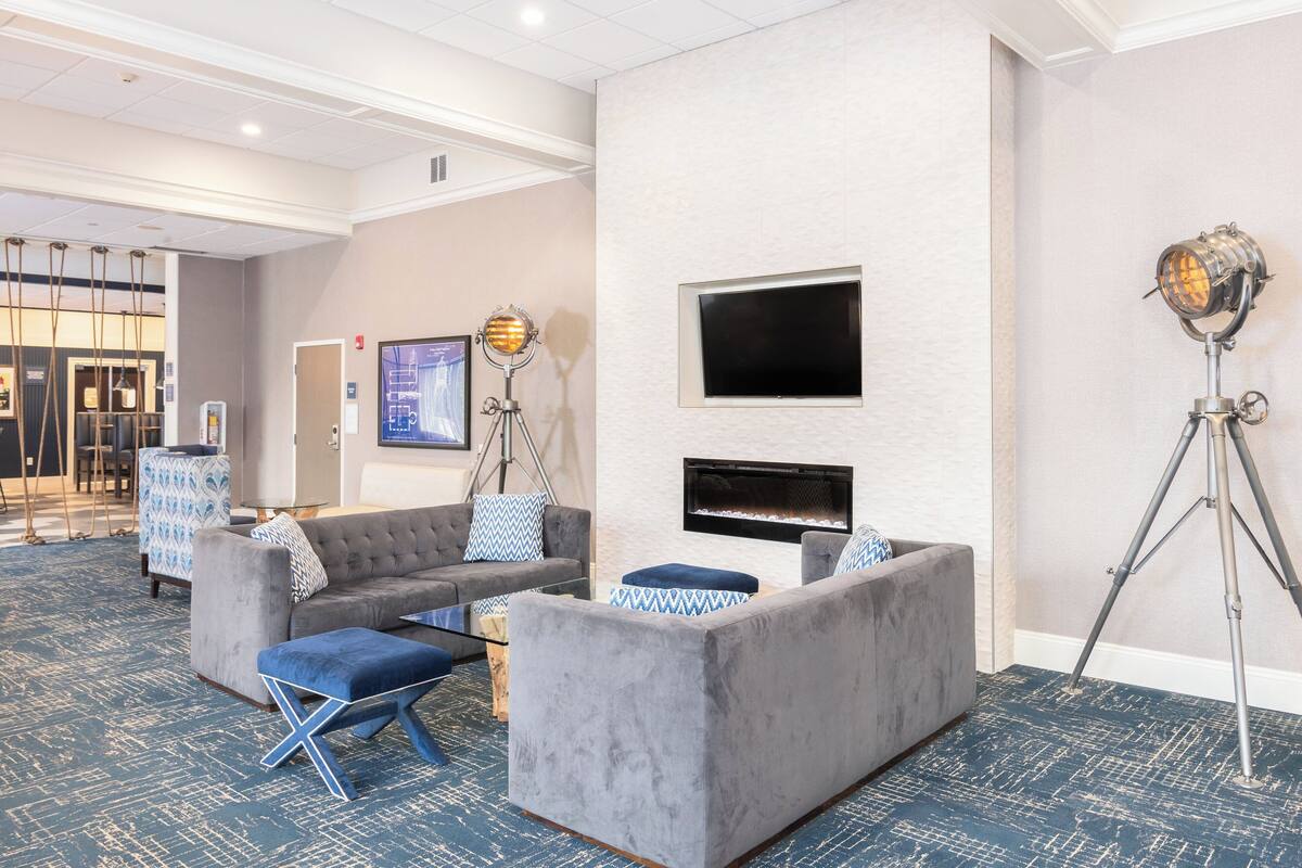 Un moderno vestíbulo de hotel con un sofá gris, un reposapiés azul y un televisor de pantalla plana montado en la pared.