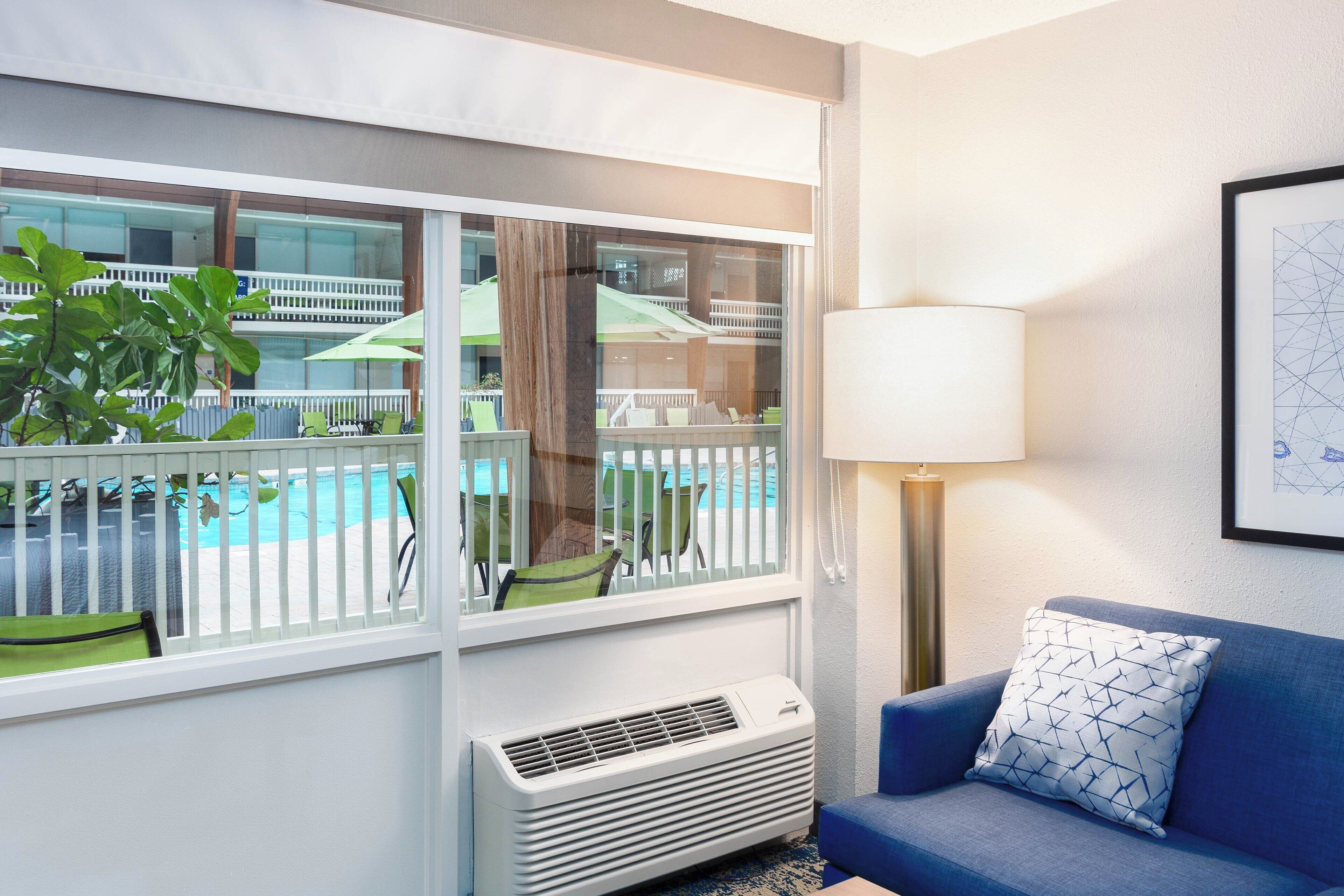 A hotel room with a large window, a blue sofa, a white air conditioner, and a lamp.