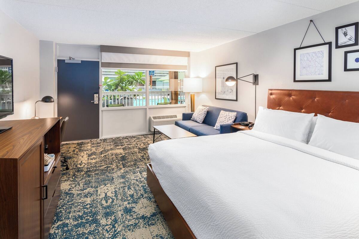 Habitación de hotel con cama, escritorio, sofá, ventana con vista a una piscina y cuadros en la pared.