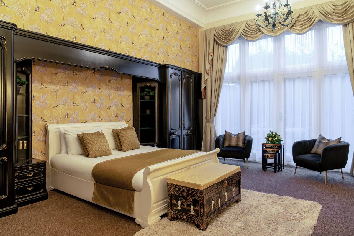 Une chambre à coucher avec un grand lit, une table de chevet, une commode, deux fauteuils et une petite table sur laquelle est posée une plante.