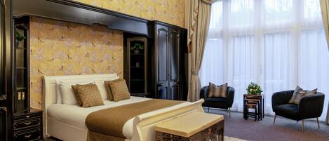 A bedroom with a large bed, a nightstand, a chest of drawers, two armchairs, and a small table with a plant.