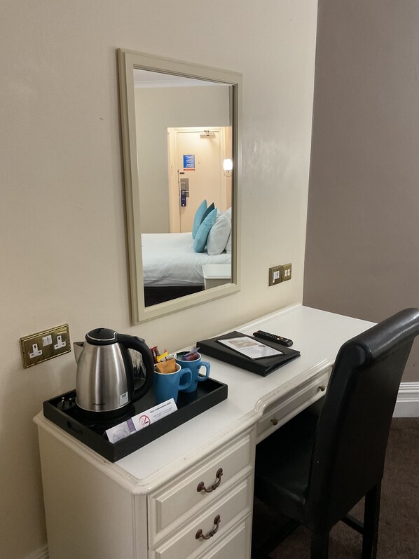 A hotel room with a white desk, a black chair, a kettle, a tray with a mug and a remote control, and a mirror reflecting the room.
