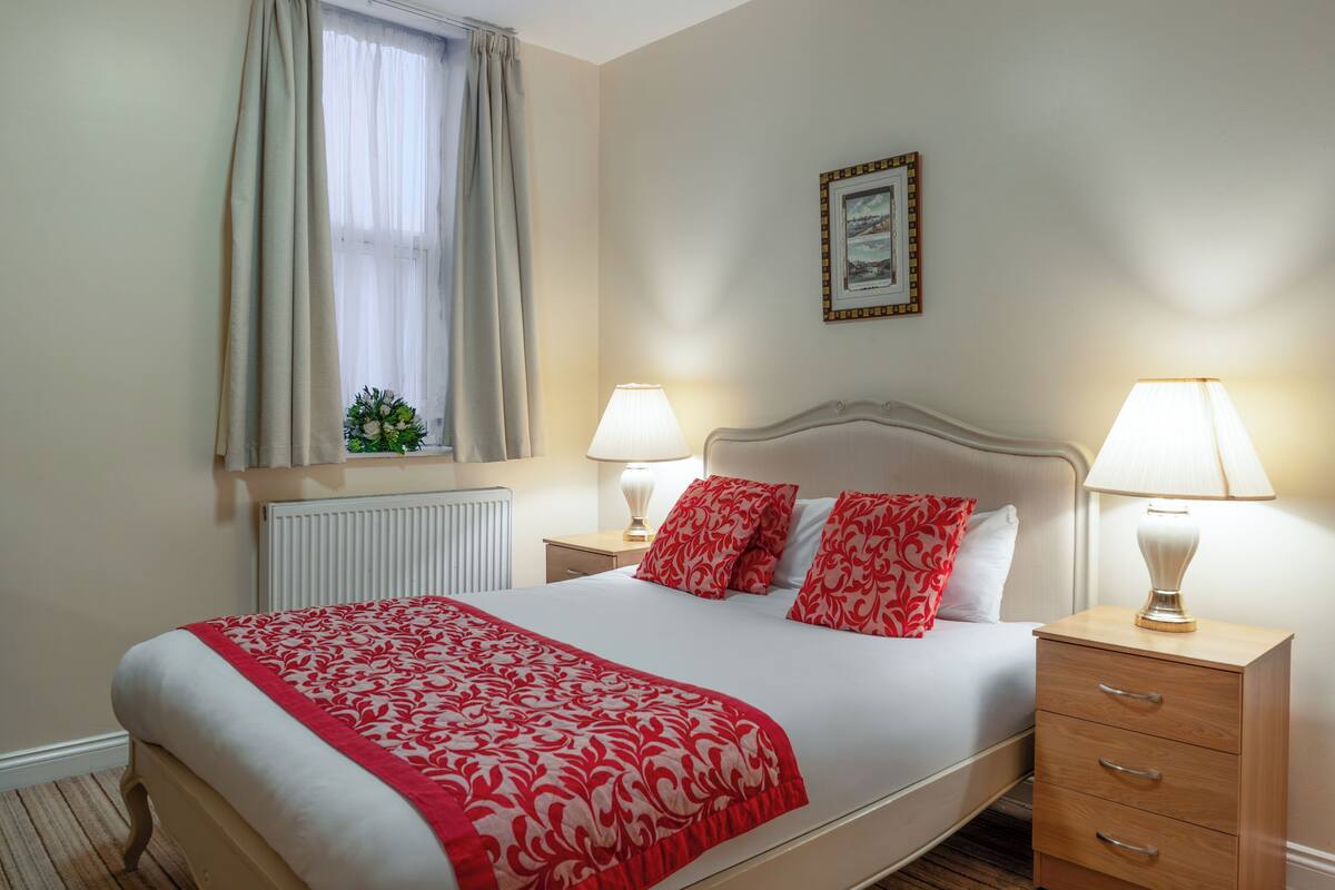 Un lit bien fait, recouvert d’une couvre-lit à motifs rouges et blancs, deux lampes de chevet, une table de chevet avec des tiroirs, une fenêtre avec des rideaux et un tableau encadré accroché au mur.