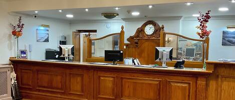 Un área de recepción con un mostrador de madera, dos monitores, un reloj y un espejo decorativo.