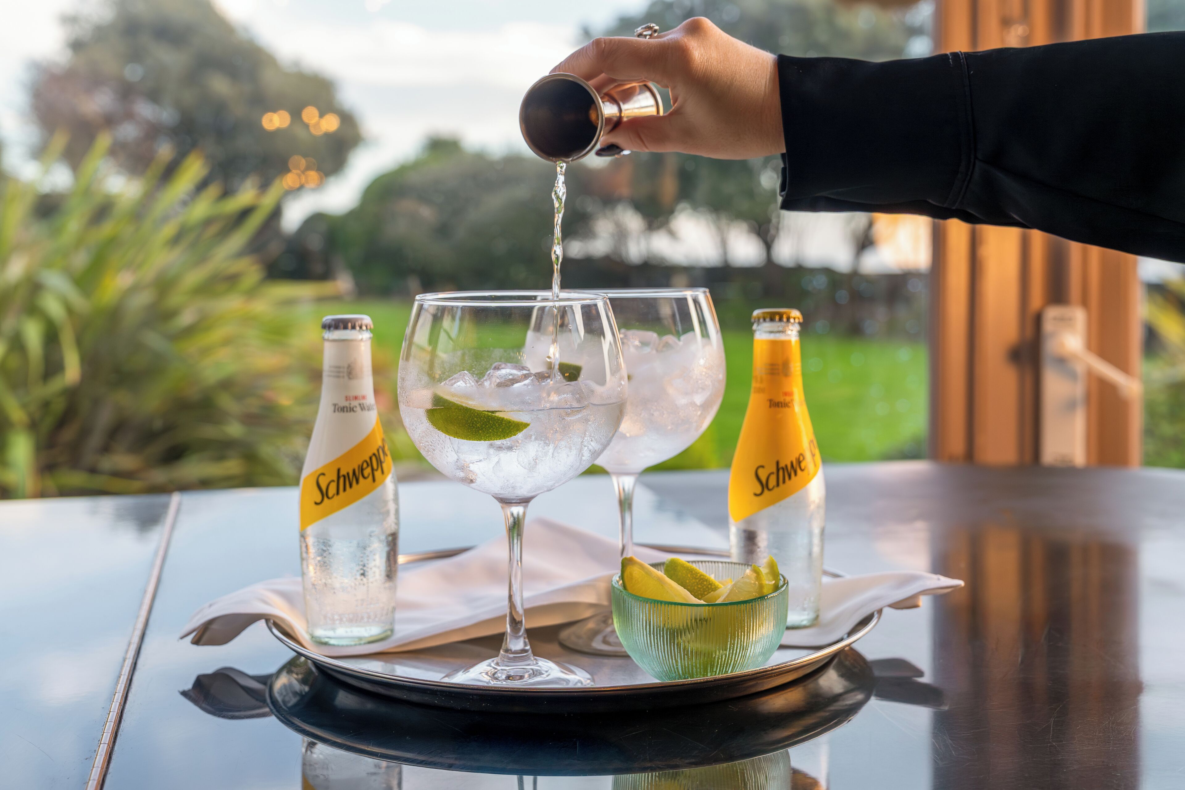 A hand pouring a drink into a glass with ice, two bottles of Schweppes tonic water, and a bowl of lime slices on a table with a view of greenery.