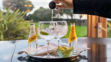 A hand pouring a drink into a glass with ice, two bottles of Schweppes tonic water, and a bowl of lime slices on a table with a view of greenery.
