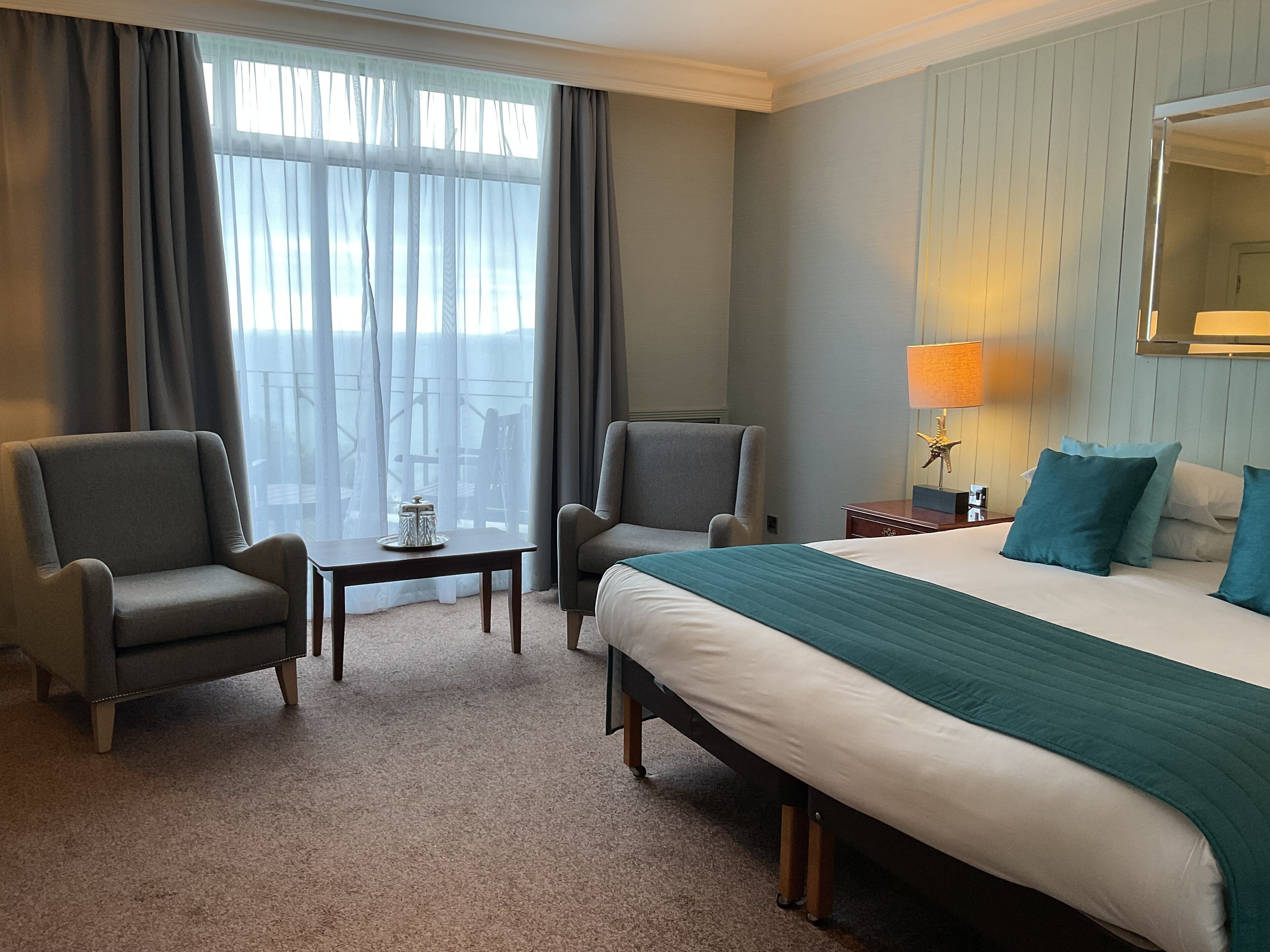 A hotel room with a bed, two armchairs, a small table, a lamp, and a view through the window.