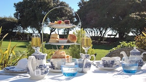 A three-tiered tea service set with various pastries and sandwiches, a teapot, cups, and a jug, all arranged on a table outdoors.