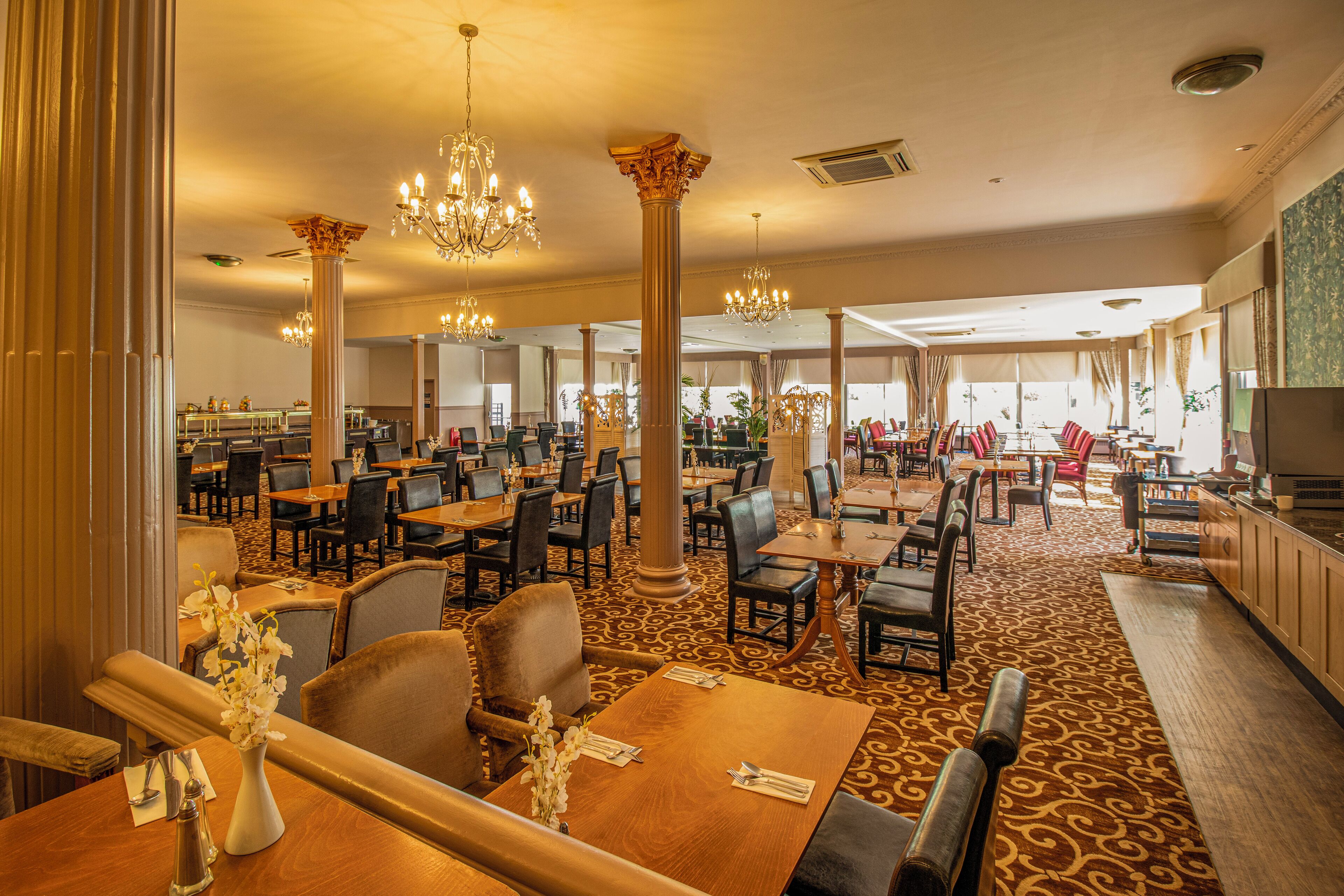 Un amplio comedor de hotel con numerosas mesas y sillas, candelabros elaborados y ventanas grandes que dejan entrar luz natural.