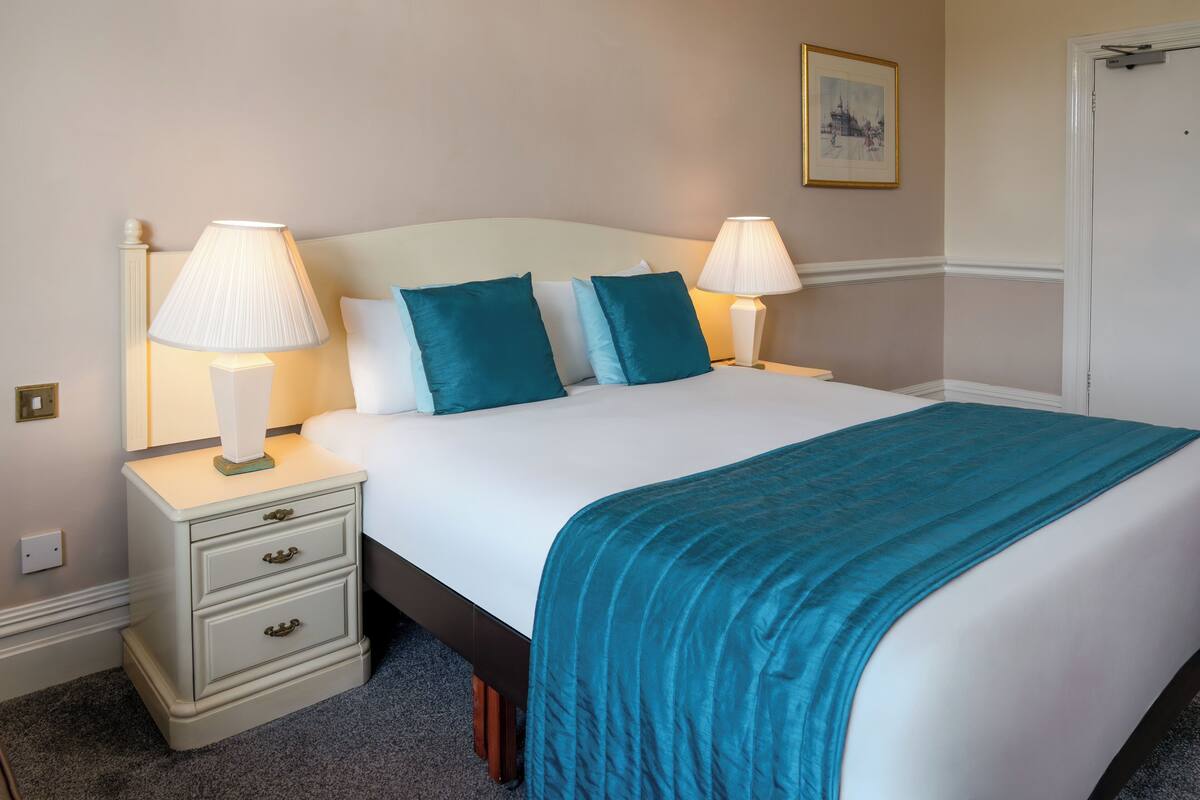 Un lit bien fait, recouvert d’une couvre-lit turquoise, avec deux lampes de chevet et une petite table de nuit dotée de tiroirs.