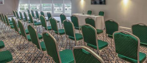 Un salón de conferencias con filas de sillas verdes, una mesa con mantel blanco y ventanas amplias con vistas a la vegetación.