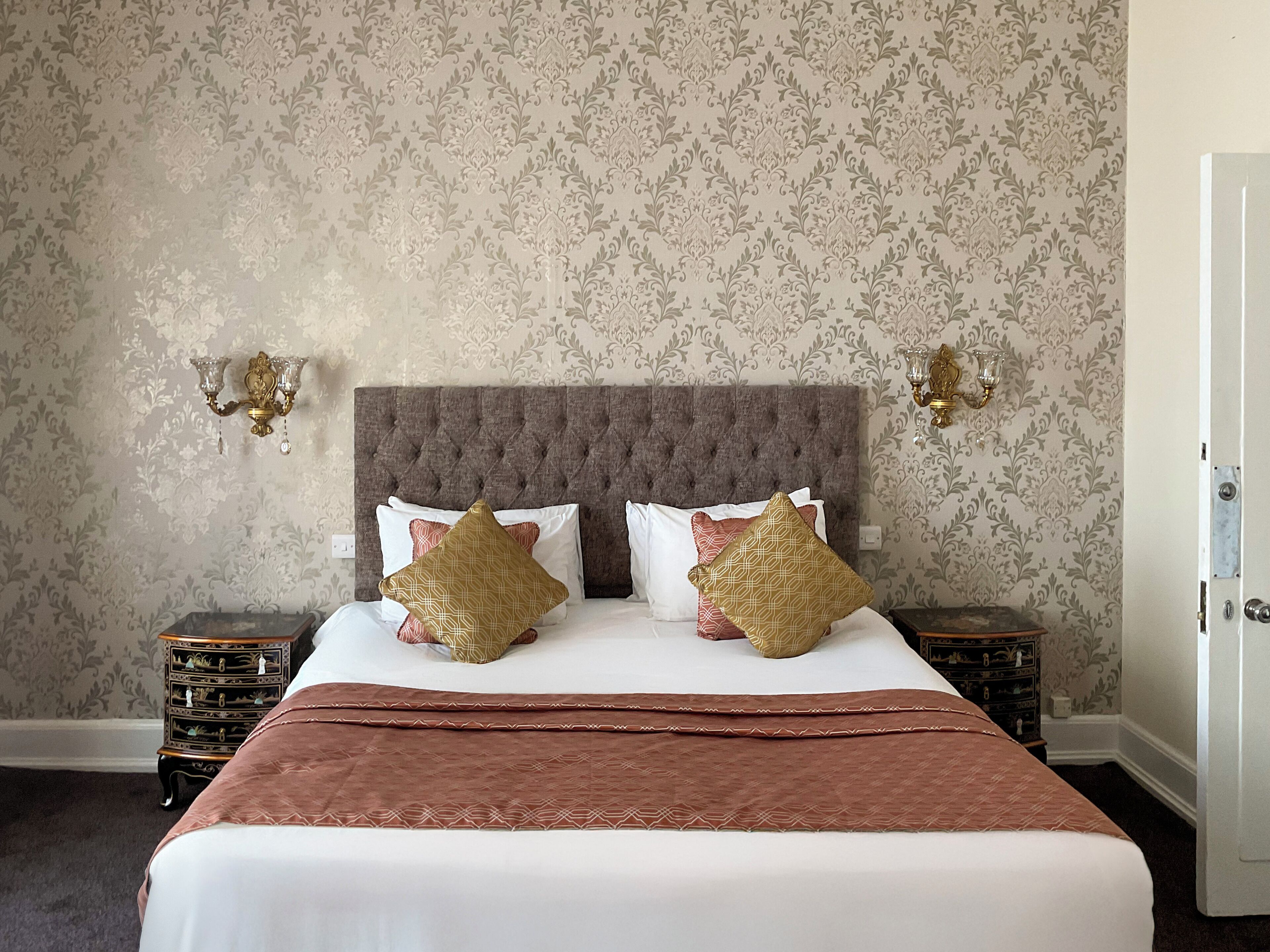 A neatly made bed with a brown headboard, two bedside tables with lamps, and patterned wallpaper.