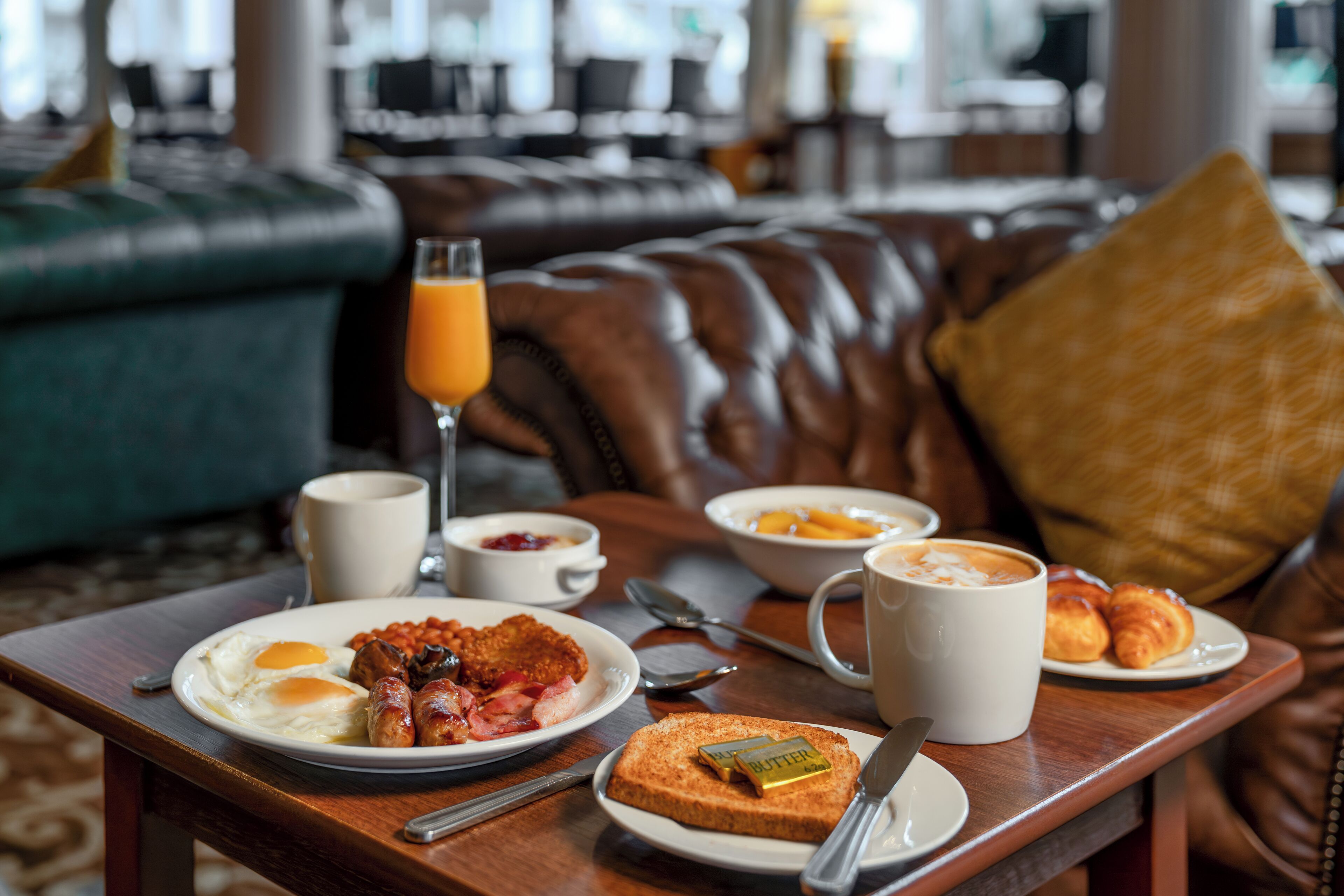 A breakfast tray with eggs, bacon, sausages, toast, and a glass of orange juice on a wooden table in a hotel lobby.