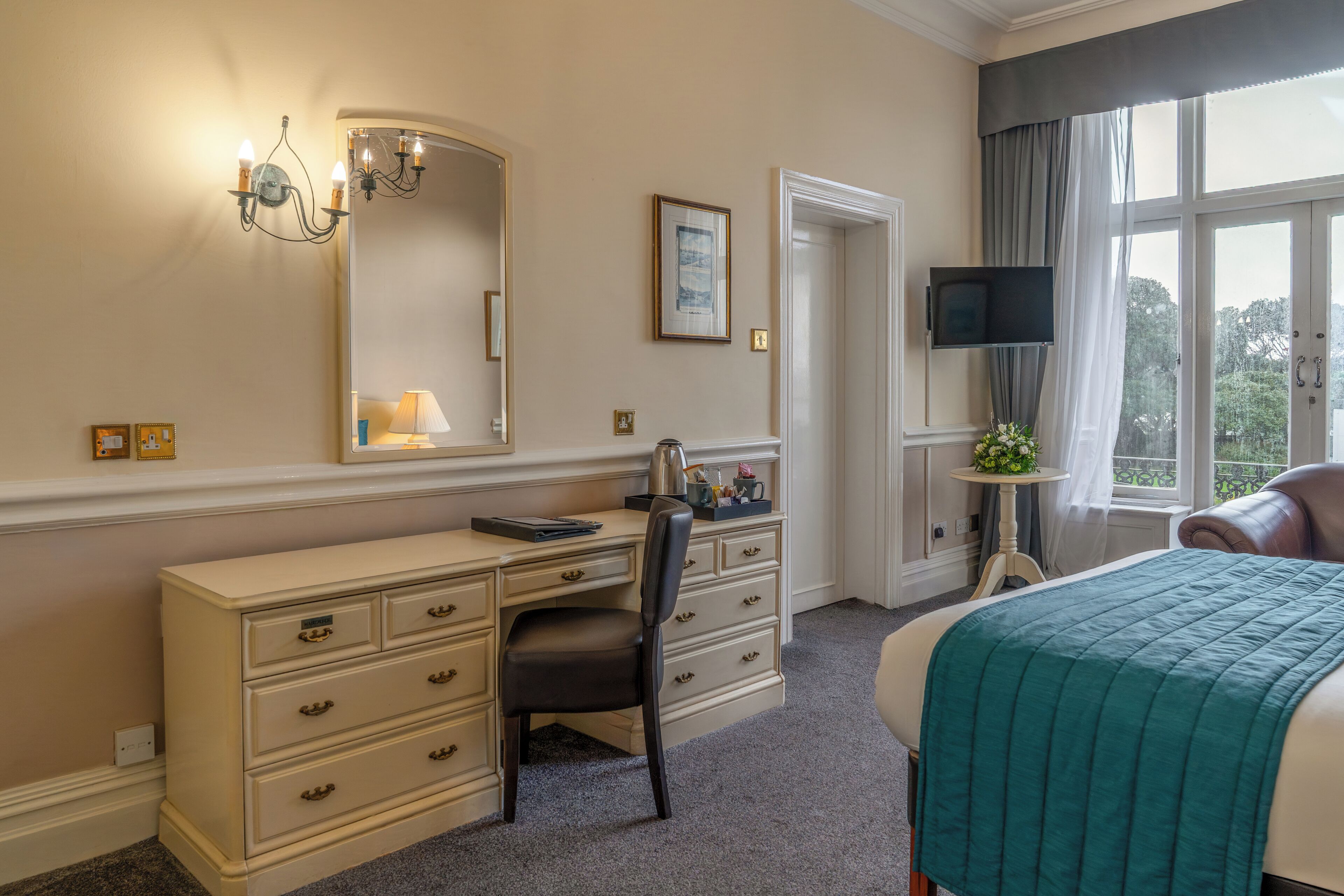 A hotel room with a bed, a desk with drawers, a chair, a television, a window with curtains, and a mirror.