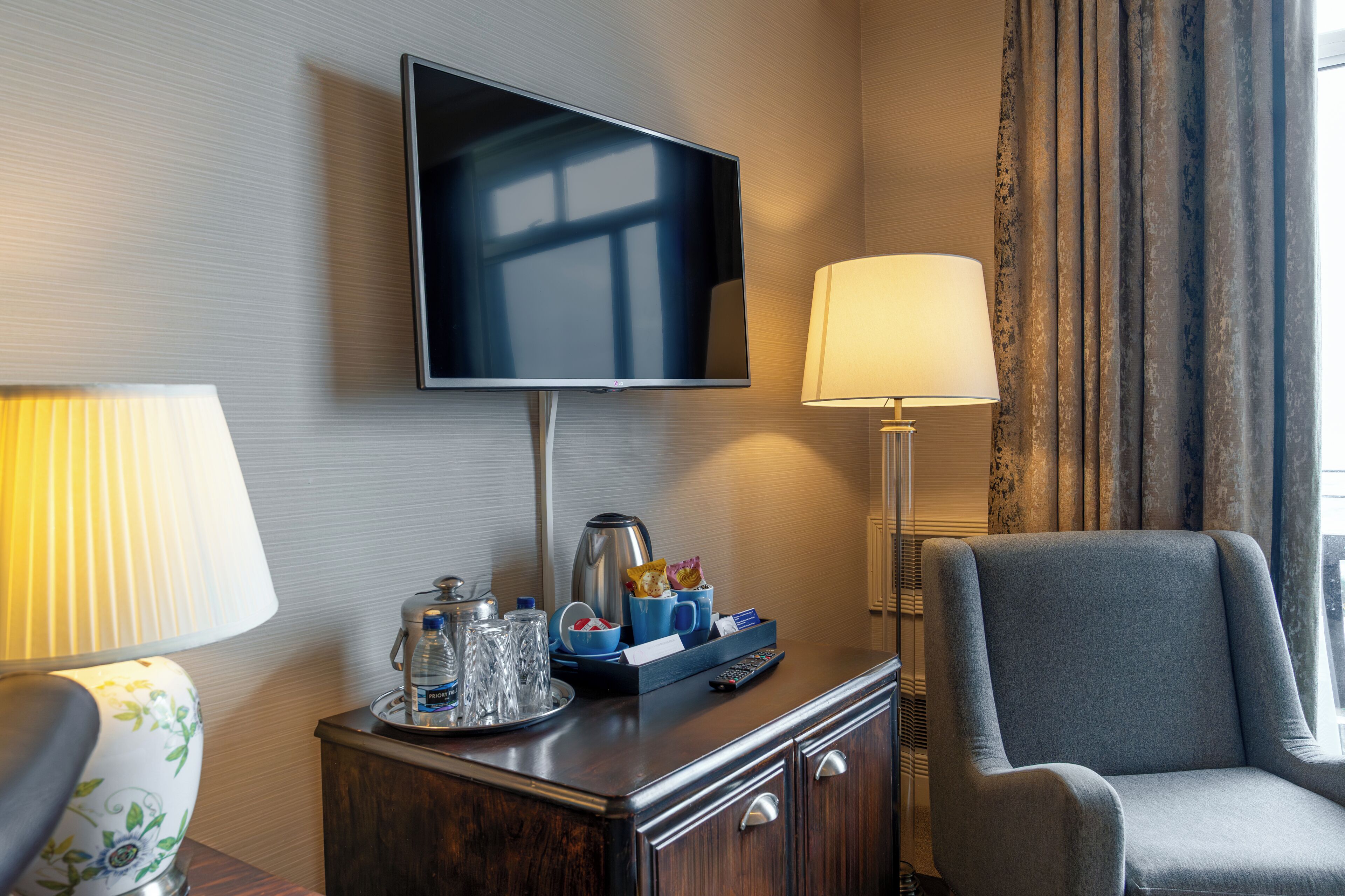 A hotel room with a flat-screen TV mounted on the wall, a bedside table with a lamp, a chair, and a tray with a coffee pot and other items.