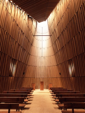 A church interior with wooden pews arranged in rows, a central aisle, and a high ceiling with vertical wooden slats.