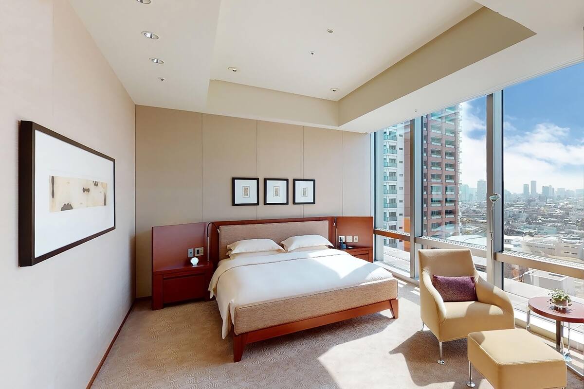 A modern hotel room with a large bed, a chair, a small table, and a city view through large windows.