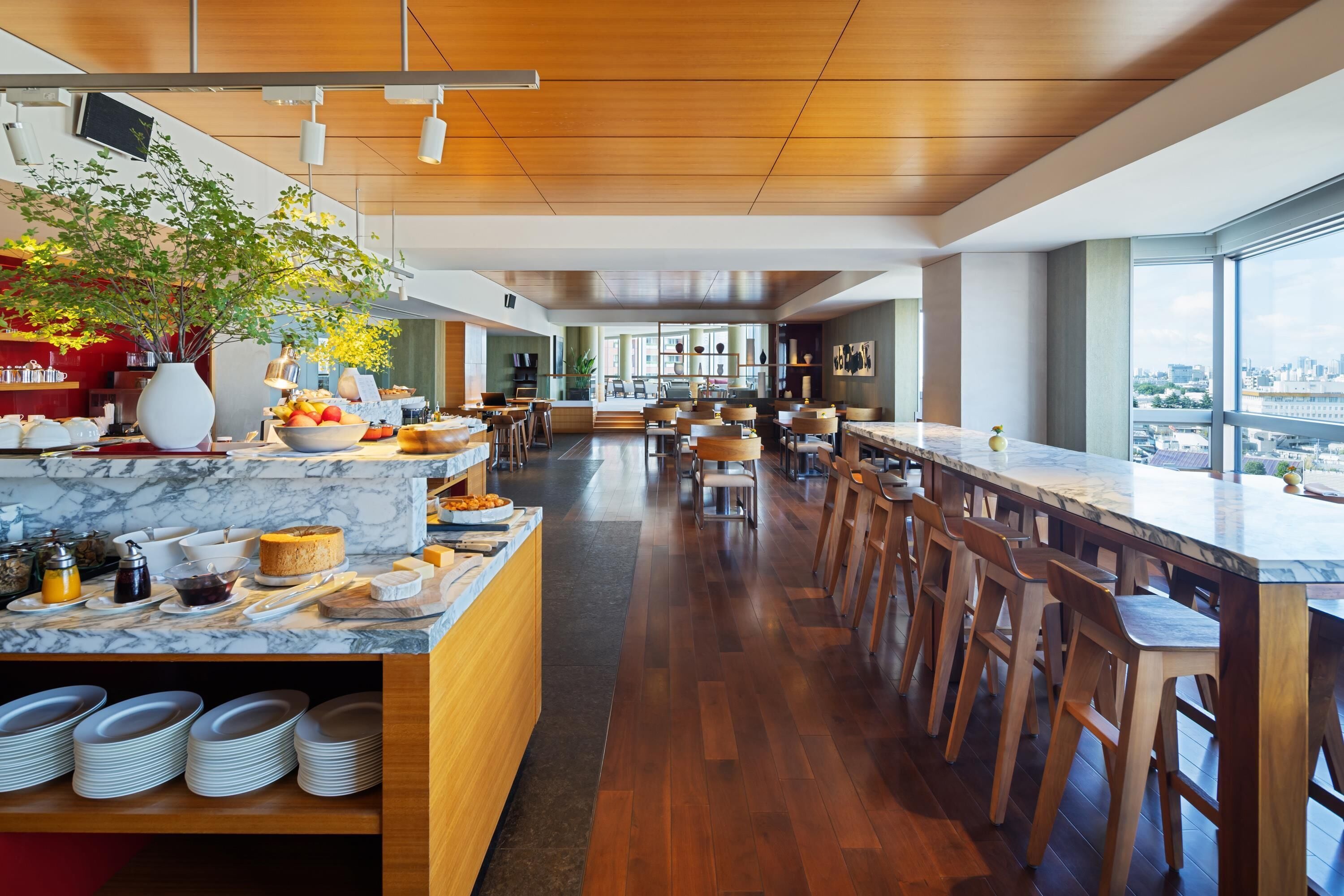 A modern restaurant with a long counter, wooden chairs, and a view of the city.