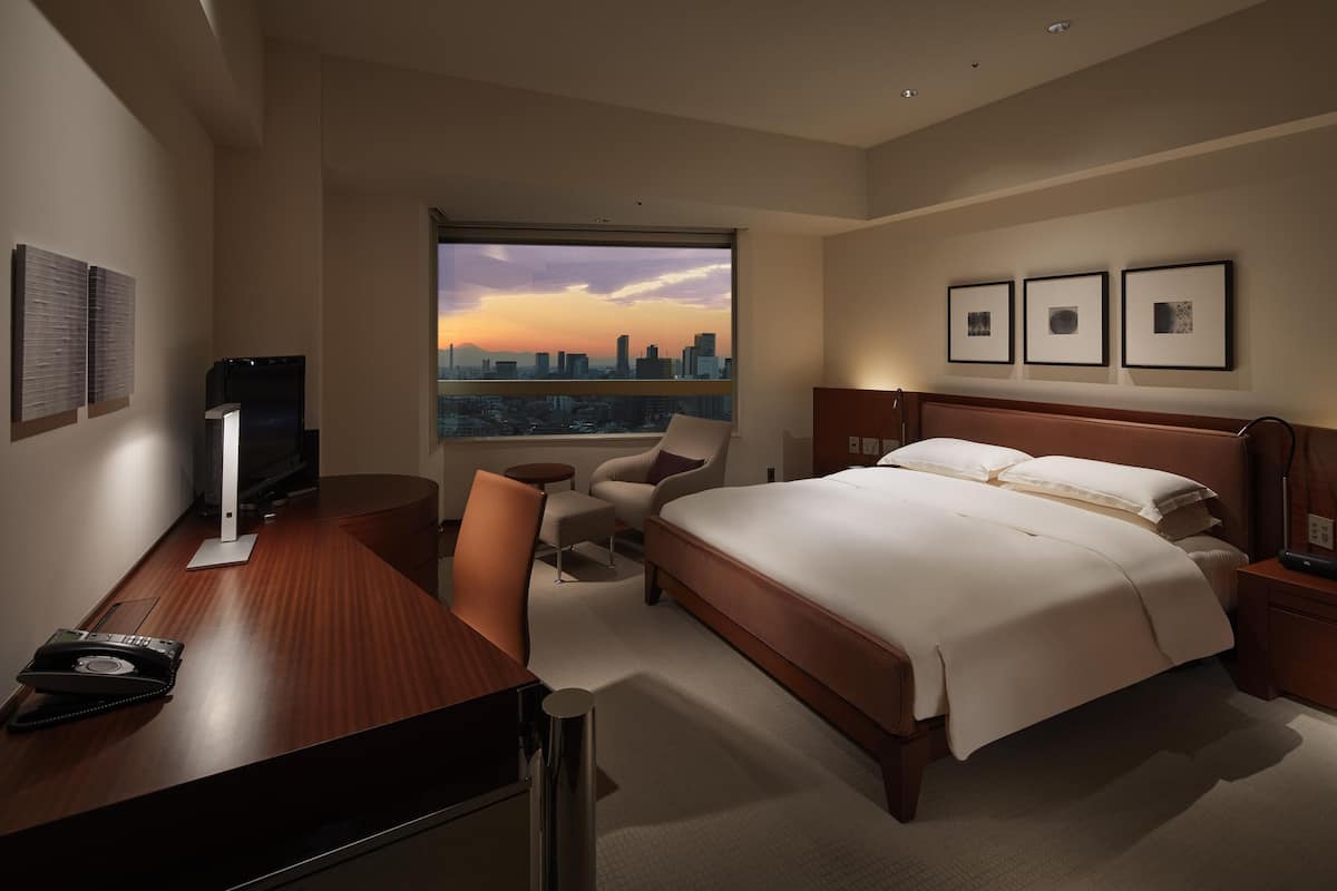 A hotel room with a large bed, a desk with a telephone, a chair, a small table, and a city view through a large window.