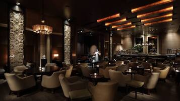 A dimly lit restaurant with a bar area, featuring a chandelier, wooden tables, and cushioned chairs.