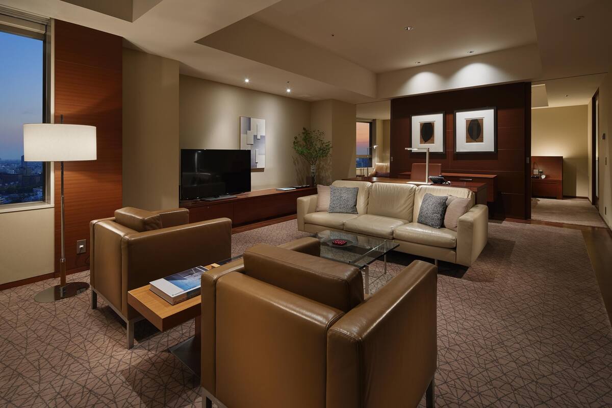 A modern hotel room with leather sofas, a glass coffee table, a flat-screen TV, and a city view.