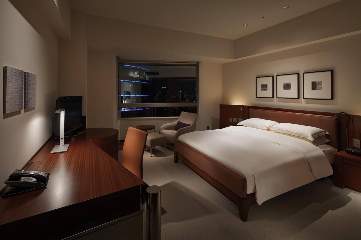 A hotel room with a bed, desk, chair, and a large window with a city view.
