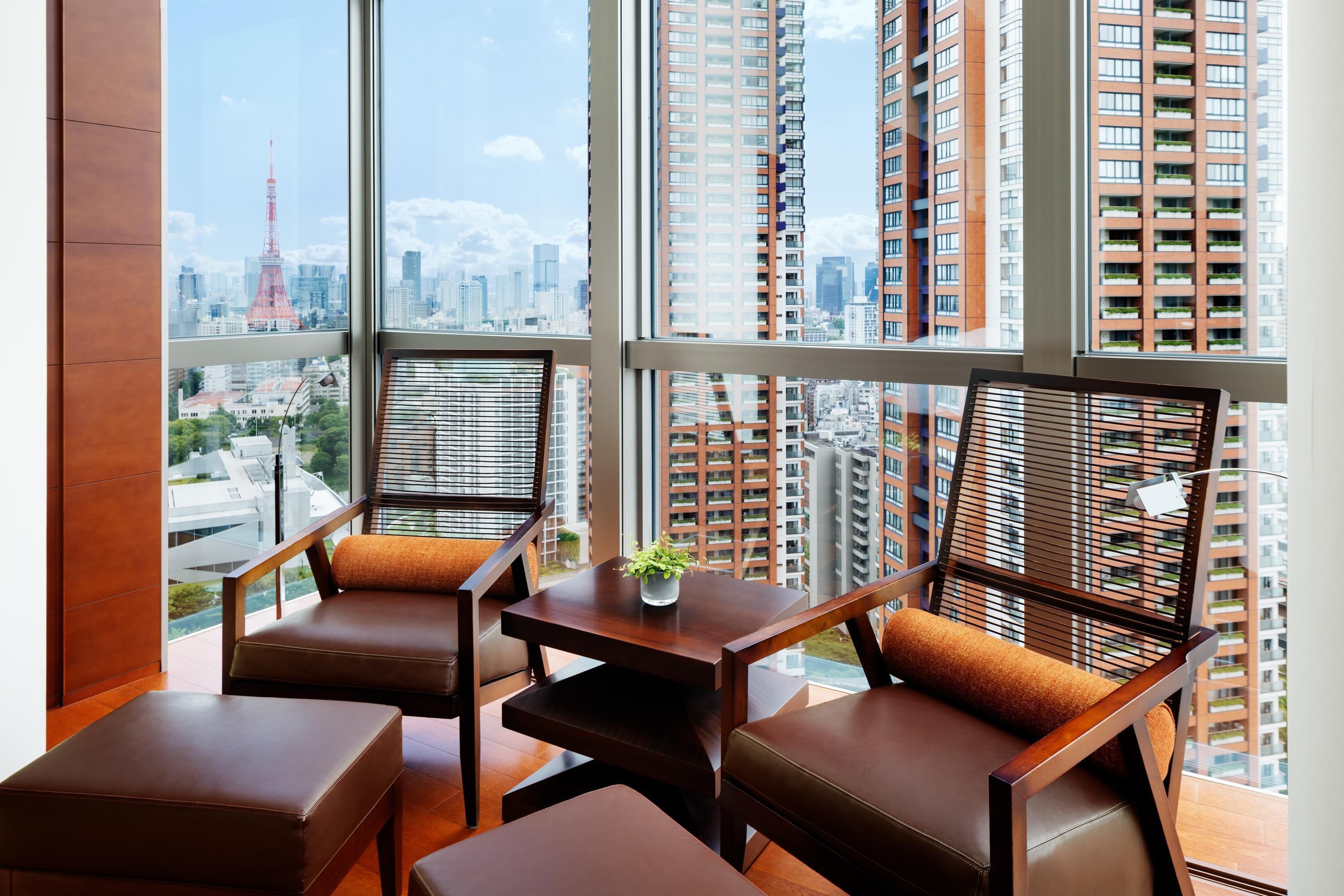 A modern room with a large window, two chairs, a small table with a plant, and a view of high-rise buildings and a tower.