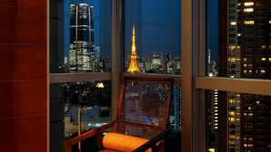 A chair with a cushion, a window with blinds, and a cityscape view including a tall illuminated building.