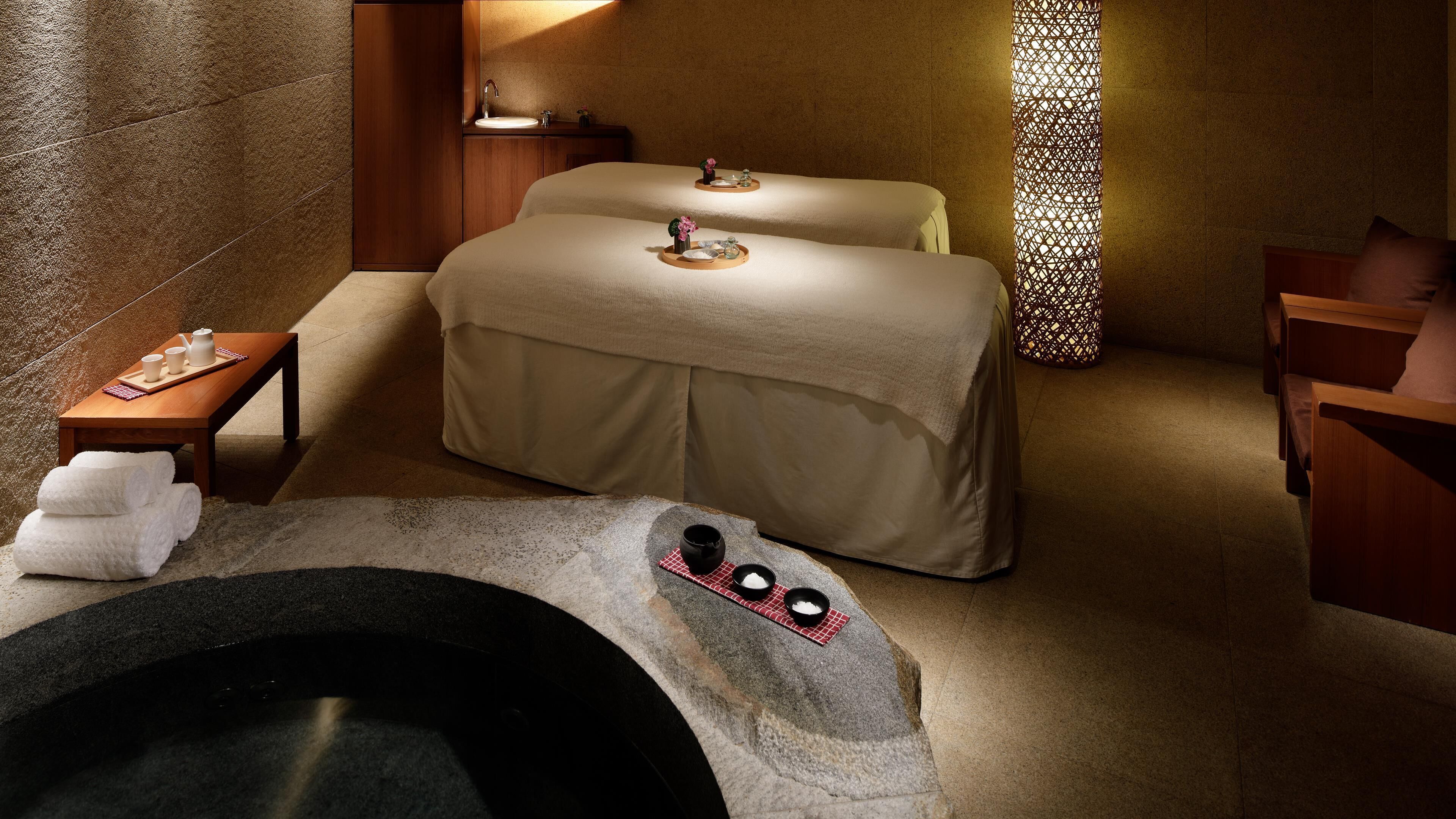 Ein Spa-Raum mit Massageliege, einem kleinen Tisch mit Handtüchern und Tablett, einem Steingefäß und einer dekorativen Wandverkleidung.